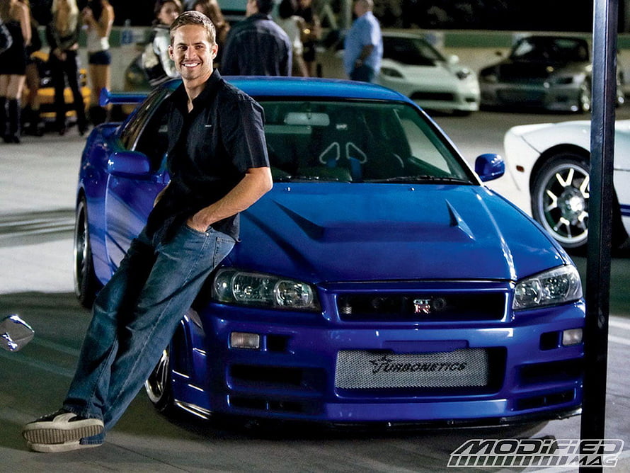 Paul Walker leaning against a blue Nissan Skyline GT-R R34 at a car meet HD wallpaper