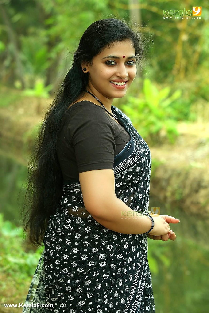 Anu Sithara in a black and white patterned saree with long dark hair in a lush green outdoor setting HD phone wallpaper