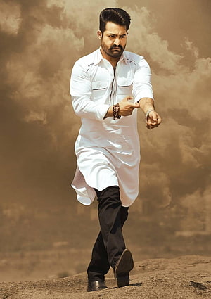 JR NTR in a white kurta and black trousers walking on a rocky terrain under a dramatic cloudy sky HD phone wallpaper