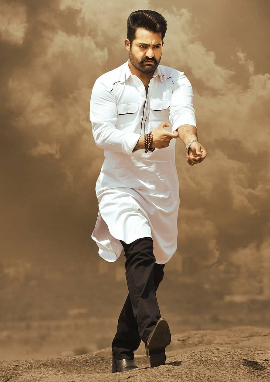 JR NTR in a white kurta and black trousers walking on a rocky terrain under a dramatic cloudy sky HD phone wallpaper